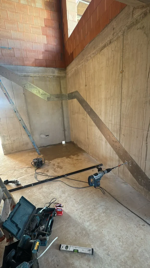 Metallschablone zur Treppenmontage an einer Betonwand befestigt, Bohrmaschine im Einsatz, Werkzeugkoffer und Wasserwaage im Vordergrund. Vorbereitung für die Treppeninstallation im Neubau.