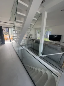 Detailaufnahme einer modernen Mittelholmtreppe mit Glasgeländer und weißen Stufen. Die Treppe führt in ein helles Wohnzimmer mit grauer Couch. Maßgefertigte Treppen für Ihr individuelles Zuhause.