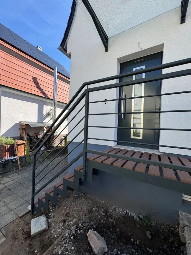Außentreppe mit Stahlgeländer und Holzstufen führt zur Haustür. Maßgefertigte Treppe im modernen Design für den Eingangsbereich. Detailaufnahme der Treppenkonstruktion mit Fokus auf die hochwertige Verarbeitung.