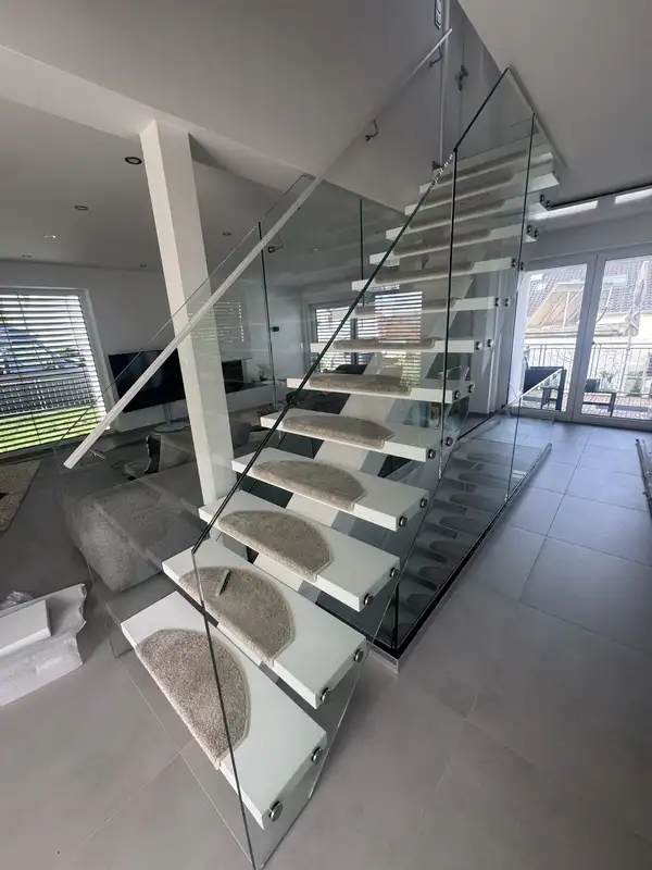 Moderne Mittelholmtreppe mit weißen Stufen und beigen Stufenmatten. Die Treppe ist mit Glasgeländer ausgestattet und führt in ein helles Wohnzimmer. Maßgefertigte Treppen für Ihr Zuhause.
