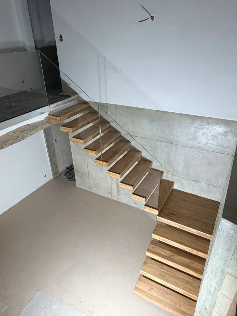 Moderne freitragende Holztreppe mit Glasgeländer vor einer Betonwand. Die Treppe ist aus hellem Holz gefertigt und führt in ein offenes Geschoss. Treppen nach Maß für Ihr Zuhause.